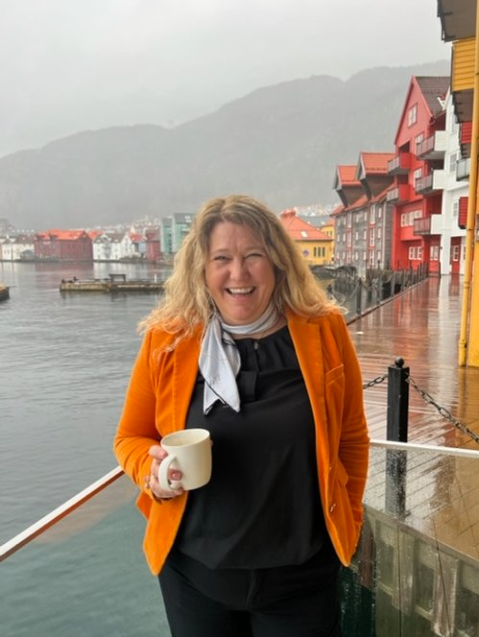 Monica ler på brygge mens hun holder en kaffekopp foran sjø
