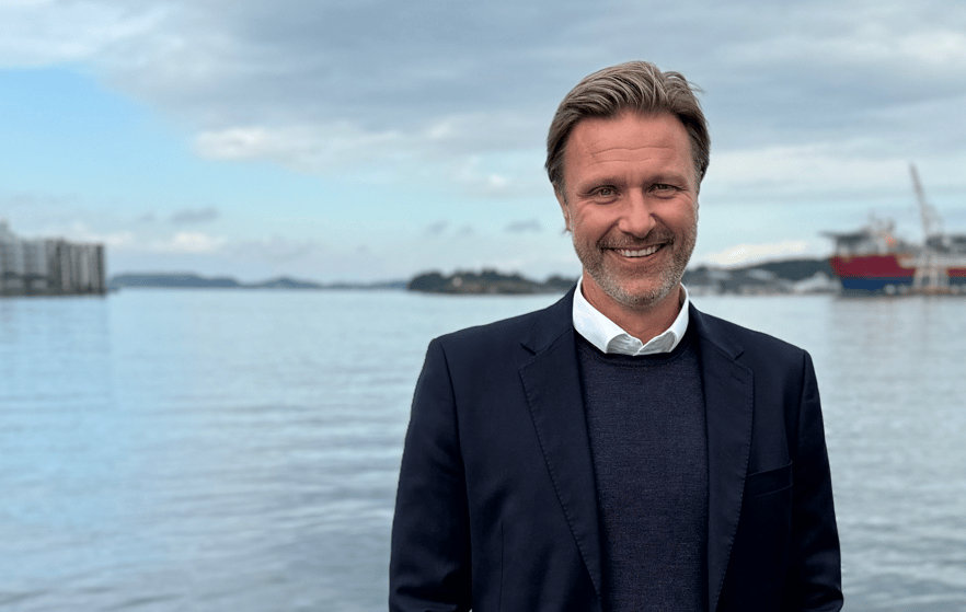 Mann (Trond) i dress smiler foran blått hav